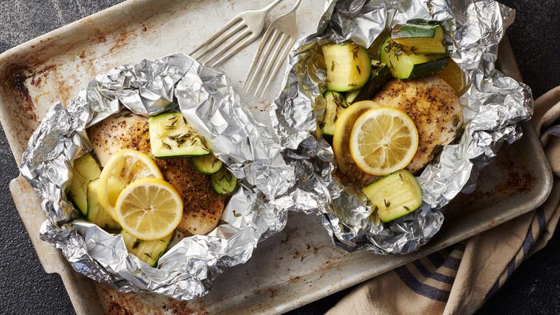 Aerial view of two foil packs filled with seasoned chicken breasts, zucchinis, and lemon slices arranged on a cooking pan. Two forks and a blue and cream-colored napkin are placed nearby.